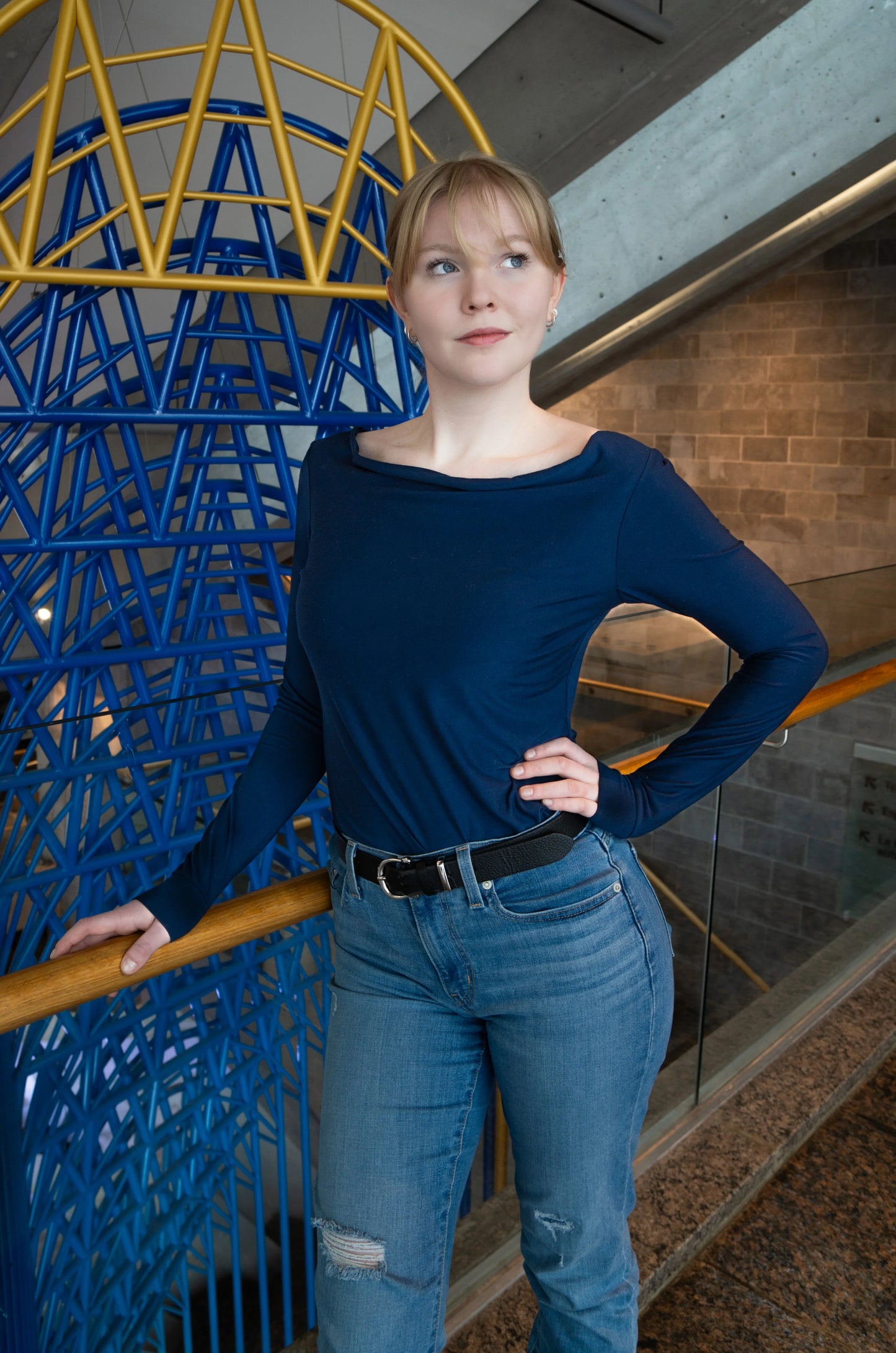 A woman wearing the Drapé Top by Karkass in Navy, a long sleeved rayon jersey top with a draped boatneck and a slim fit. she is standing indoors in front of a colourful structure. 