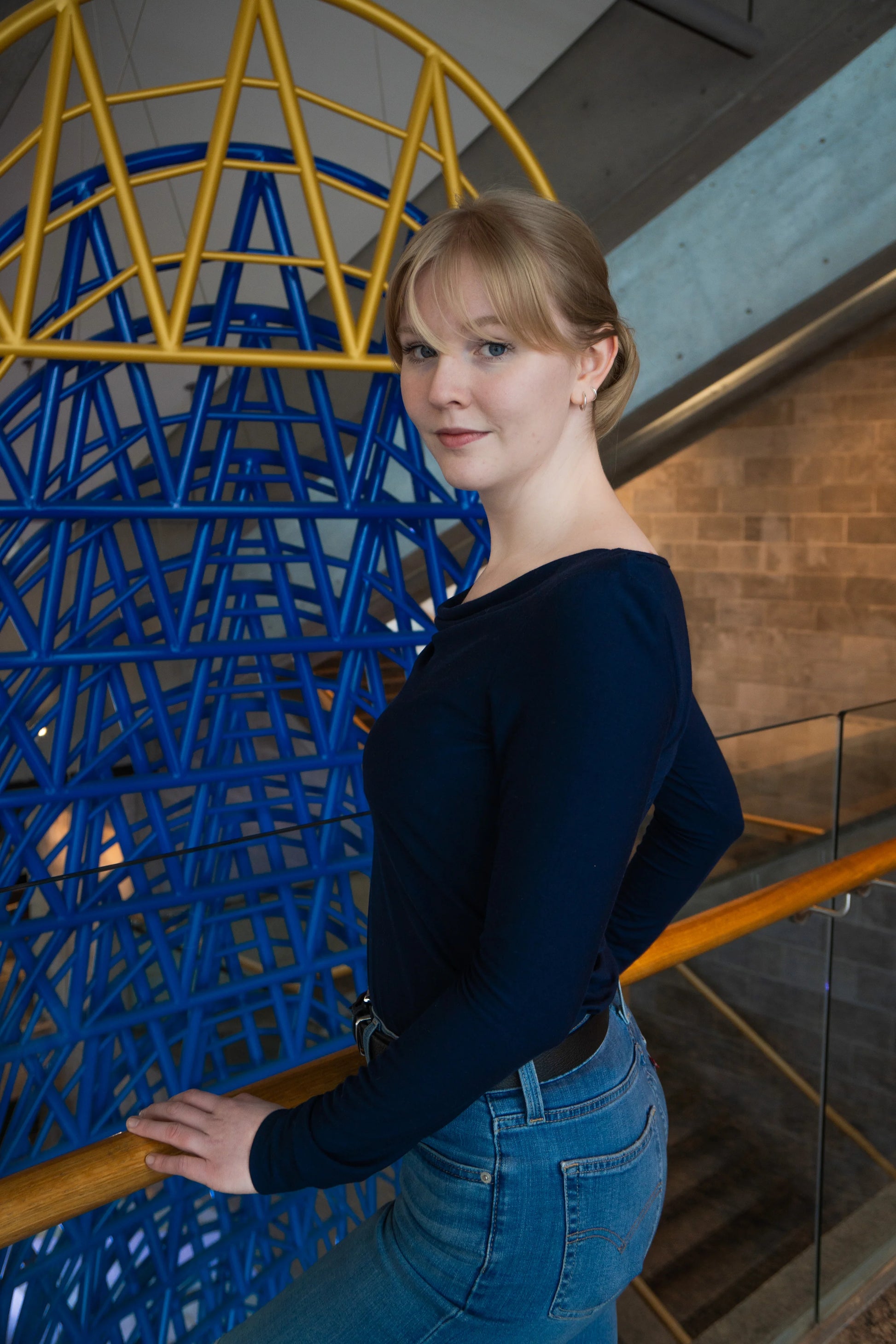 Side view of a woman wearing the Drapé Top by Karkass in Navy, a long sleeved rayon jersey top with a draped boatneck and a slim fit. she is standing indoors in front of a colourful structure. 
