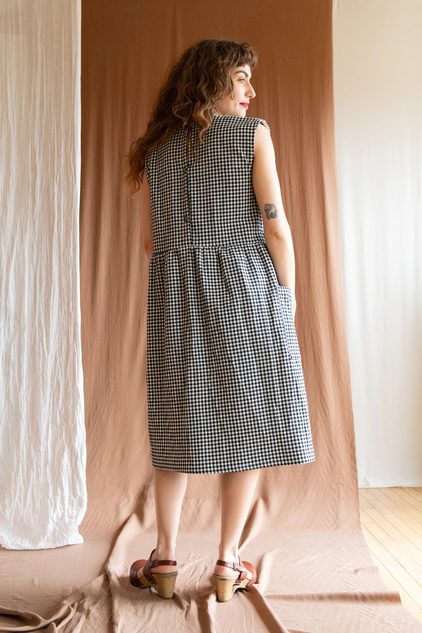 Back view of a woman wearing the Avellino Dress by Kazak in Black and White Vichy, featuring short extended sleeves, a round neck, gathered waist, full skirt that falls below the knee, a removable belt, and large patch pockets. She is standing in front of a brown curtain. 
