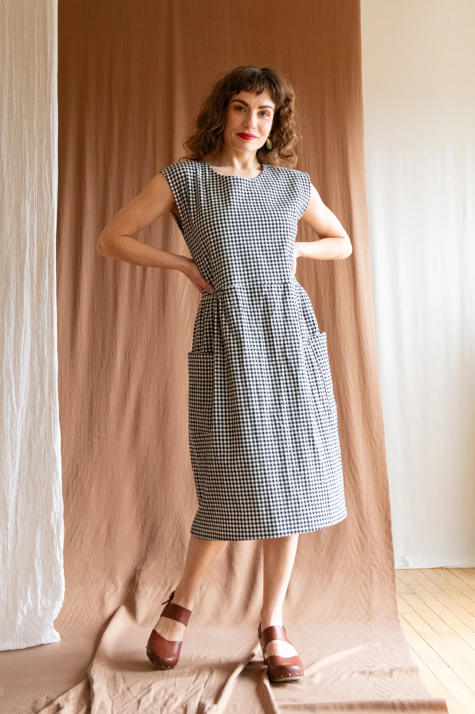 A woman wearing the Avellino Dress by Kazak in Black and White Vichy, featuring short extended sleeves, a round neck, gathered waist, full skirt that falls below the knee, a removable belt, and large patch pockets. She is standing in front of a brown curtain. 
