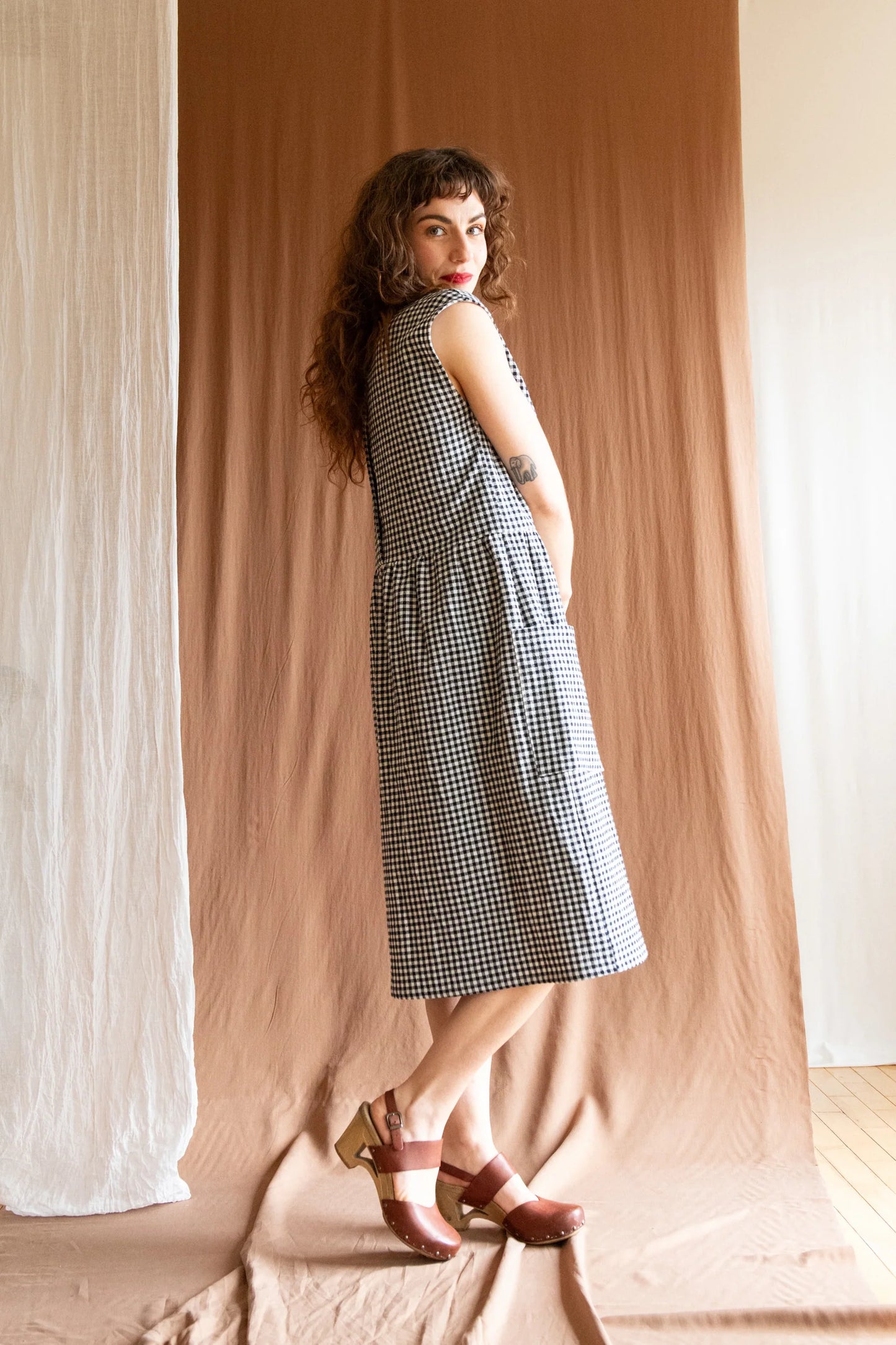 Side view of a woman wearing the Avellino Dress by Kazak in Black and White Vichy, featuring short extended sleeves, a round neck, gathered waist, full skirt that falls below the knee, a removable belt, and large patch pockets. She is standing in front of a brown curtain. 