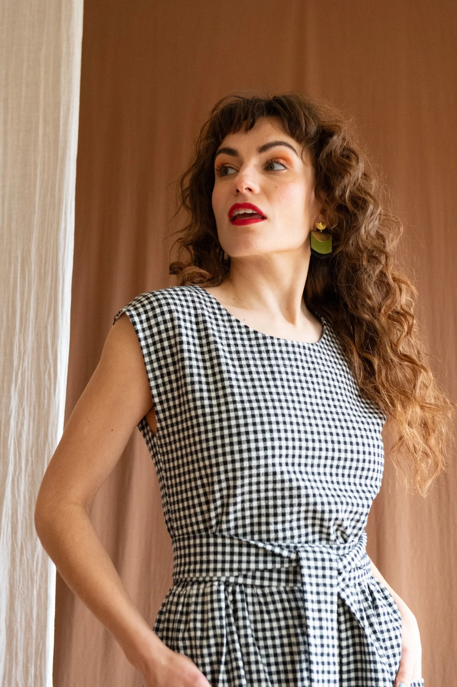 3/4 view of a woman wearing the Avellino Dress by Kazak in Black and White Vichy, featuring short extended sleeves, a round neck, gathered waist, full skirt that falls below the knee, a removable belt, and large patch pockets. She is standing in front of a brown curtain. 
