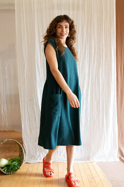 A woman wearing the Avellino Dress by Kazak in Forest, featuring short extended sleeves, a round neck, gathered waist, full skirt that falls below the knee, a removable belt, and large patch pockets. She is standing in front of a brown curtain. 