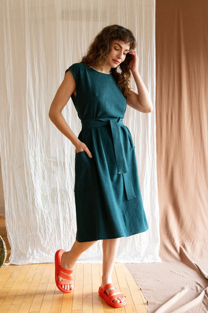 A woman wearing the Avellino Dress by Kazak in Forest, featuring short extended sleeves, a round neck, gathered waist, full skirt that falls below the knee, a removable belt, and large patch pockets. She is standing in front of a brown curtain. 