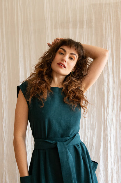 3/4 view of a woman wearing the Avellino Dress by Kazak in Forest, featuring short extended sleeves, a round neck, gathered waist, full skirt that falls below the knee, a removable belt, and large patch pockets. She is standing in front of a brown curtain. 
