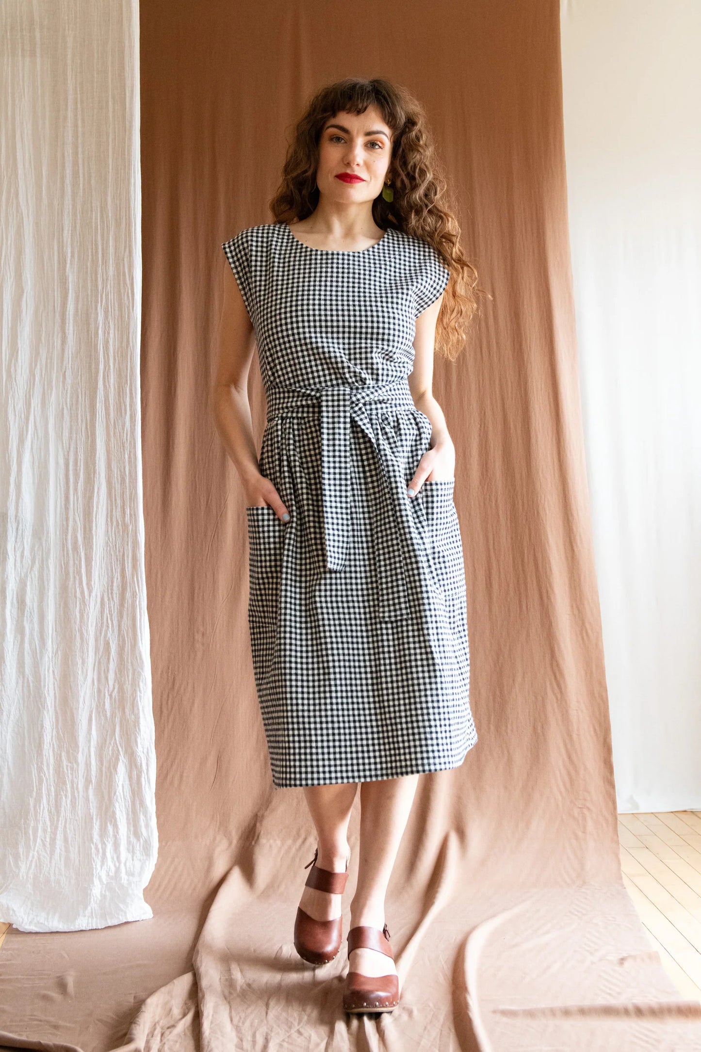 A woman wearing the Avellino Dress by Kazak in Black and White Vichy, featuring short extended sleeves, a round neck, gathered waist, full skirt that falls below the knee, a removable belt, and large patch pockets. She is standing in front of a brown curtain. 