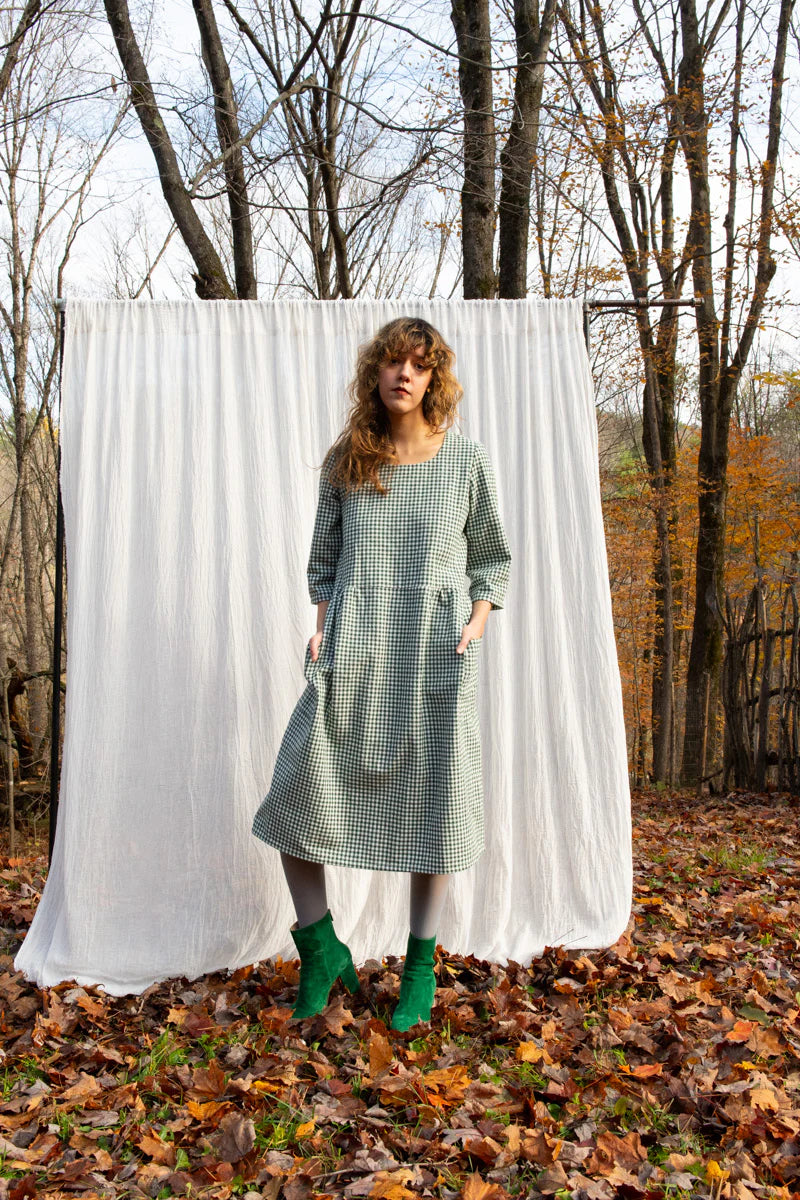 A woman wearing the Persea Dress by Kazak in Forest Vichy print, a below-the-knee length dress with 3/4 sleeves, a removable belt, and large patch pockets. She is standing in a forest against a white background. 