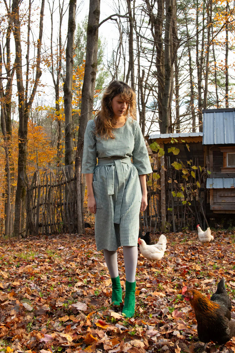 A woman wearing the Persea Dress by Kazak in Forest Vichy print, a below-the-knee length dress with 3/4 sleeves, a removable belt, and large patch pockets. She is standing in an autumn forest with chickens around her.