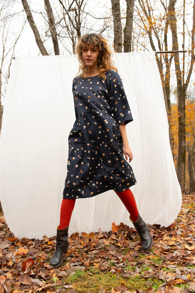 A woman wearing the Persea Dress by Kazak in Midnight Moon print, a below-the-knee length dress with 3/4 sleeves, a removable belt, and large patch pockets. She is standing in a forest in front of a white sheet. 