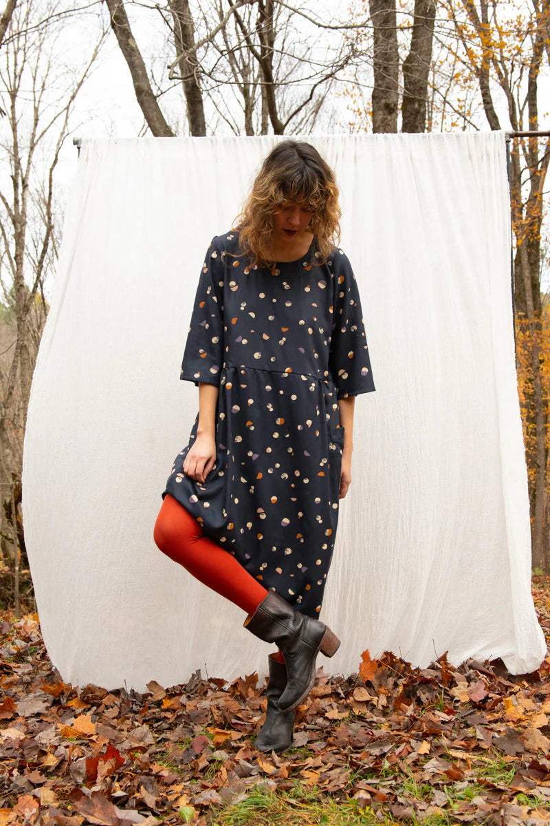 A woman wearing the Persea Dress by Kazak in Midnight Moon print, a below-the-knee length dress with 3/4 sleeves, a removable belt, and large patch pockets. She is standing in a forest in front of a white sheet. 