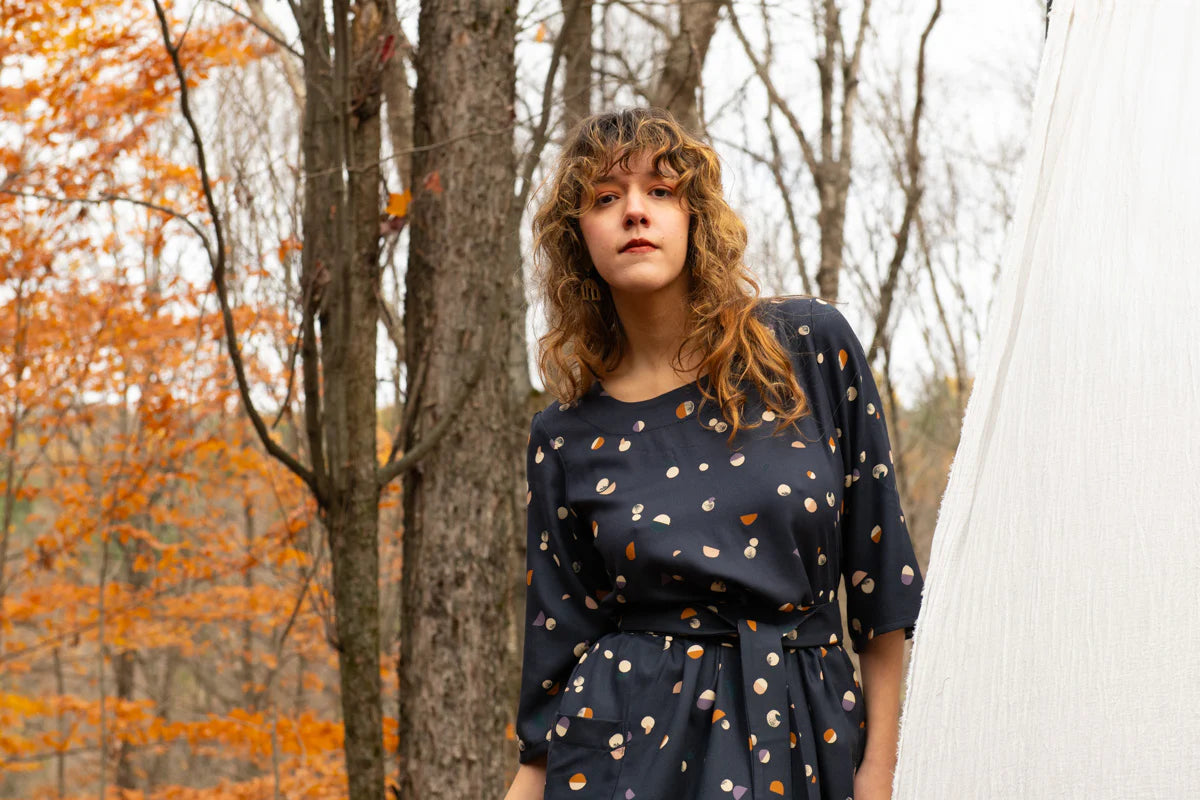 A woman wearing the Persea Dress by Kazak in Midnight Moon print, a below-the-knee length dress with 3/4 sleeves, a removable belt, and large patch pockets. She is standing in a forest.