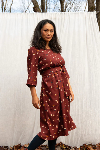 A woman wearing the Persea Dress by Kazak in Bordeaux Moon print, a below-the-knee length dress with 3/4 sleeves, a removable belt, and large patch pockets. She is standing in an autumn forest in front of a white curtain. 