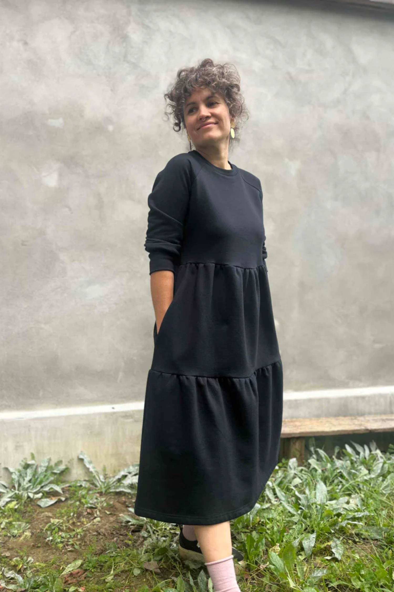 A woman wearing the Simone Dress by Kazak in Black, made from organic cotton and featuring a round neck, 3/4 raglan sleeves, and a tiered midi-length skirt. She is standing outside on front of a grey wall. 