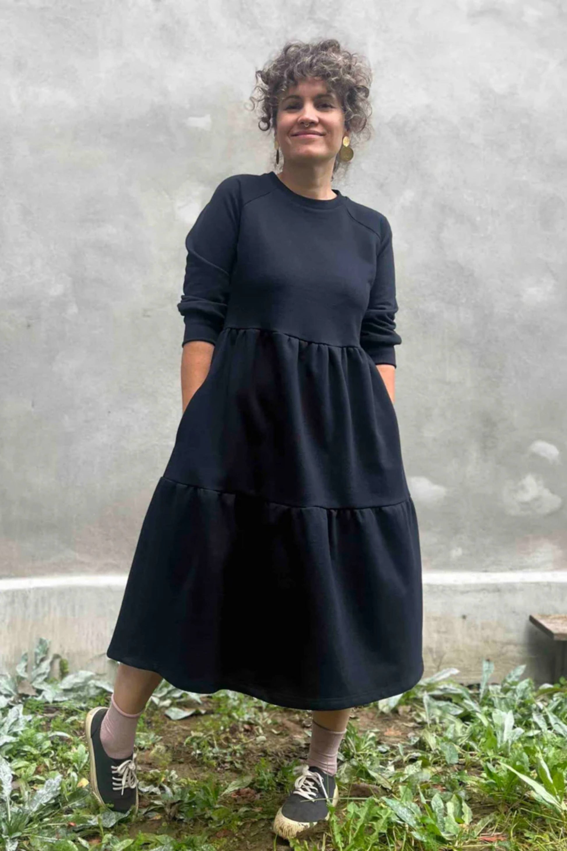 A woman wearing the Simone Dress by Kazak in Black, made from organic cotton and featuring a round neck, 3/4 raglan sleeves, and a tiered midi-length skirt. She is standing outside on front of a grey wall. 