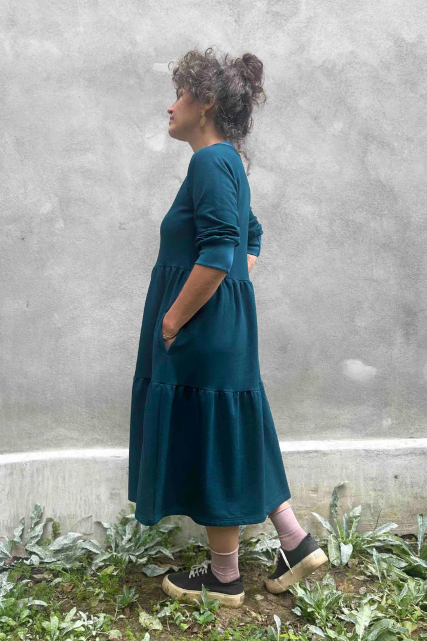 Side view of a woman wearing the Simone Dress by Kazak in Teal, made from organic cotton and featuring a round neck, 3/4 raglan sleeves, and a tiered midi-length skirt. She is standing outside on front of a grey wall. 