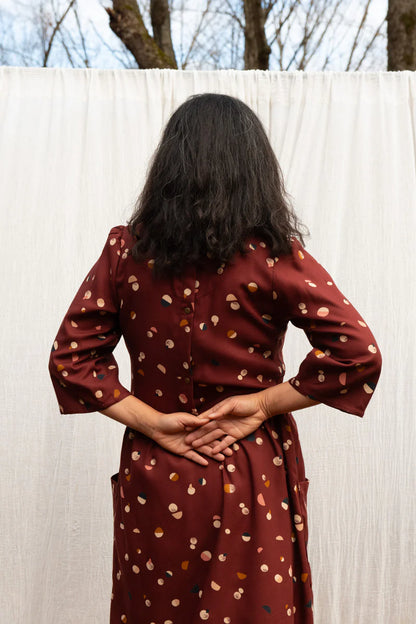 Back view of a woman wearing the Persea Dress by Kazak in Bordeaux Moon print, a below-the-knee length dress with 3/4 sleeves, a removable belt, and large patch pockets. She is standing in a forest in front of a white sheet. 