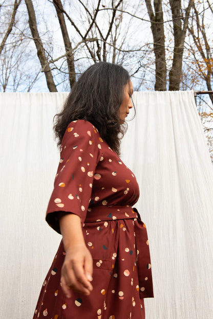 Side view of a woman wearing the Persea Dress by Kazak in Bordeaux Moon  print, a below-the-knee length dress with 3/4 sleeves, a removable belt, and large patch pockets. She is standing in a forest in front of a white sheet. 