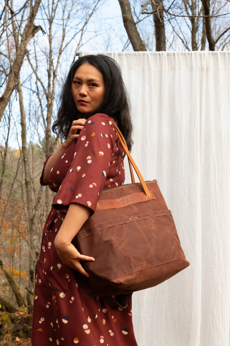 Side view of a woman wearing the Persea Dress by Kazak in Bordeaux Moon  print, a below-the-knee length dress with 3/4 sleeves, a removable belt, and large patch pockets. She is standing in a forest in front of a white sheet and holding a brown tote bag.