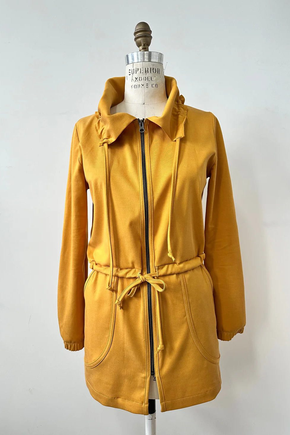 The Bronwen Jacket by Kollontai in Mustard, a mid-thigh length jacket with drawstrings at the waist and hood and large patch  pockets, is shown on a mannequin in front of a white background. 