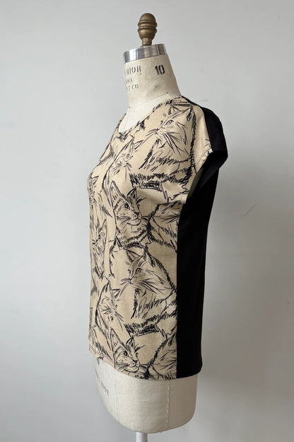 A side view of the Arty T-Shirt by Kollontai in Cats/Beige print, with a round neck, short extended sleeves, and a slight hi-low hemline, and a solid black back, is shown on a mannequin against a grey background. 