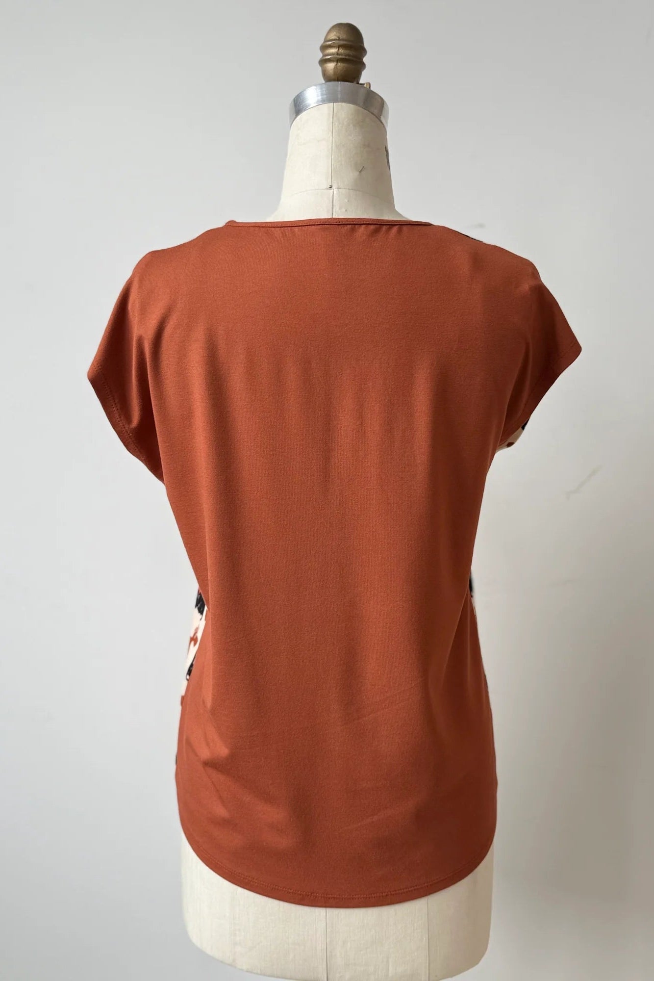 A back view of the Arty T-Shirt by Kollontai in Dogs/Rust print, with a round neck, short extended sleeves, and a slight hi-low hemline, and a solid rust back, is shown on a mannequin against a grey background. 