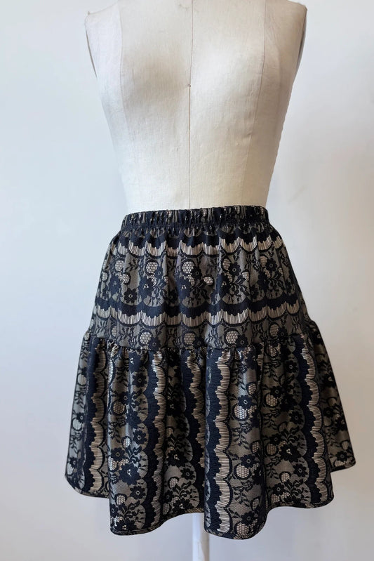 The Soleil Skirt by Kollontai, a short flounced skirt in black lace over gold satin, is shown on a mannequin  against a white background.