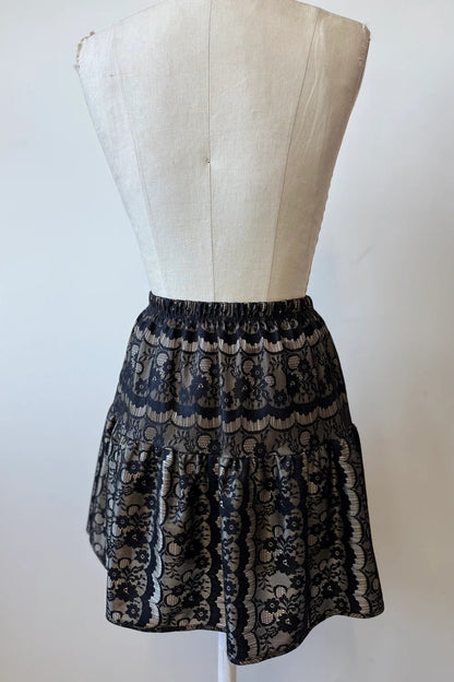 A back view of the Soleil Skirt by Kollontai, a short flounced skirt in black lace over gold satin, is shown on a mannequin  against a white background.