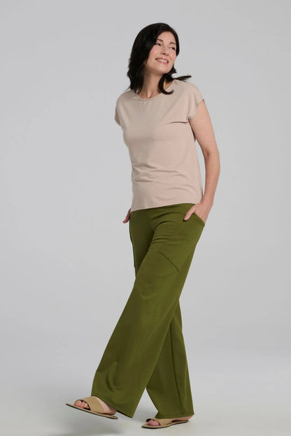 A woman wearing the Eowyn T-Shirt in Sand By Kollontai, with the spring Susan Pants by Kollontai in Granny Smith. She is standing in front of a grey background. 