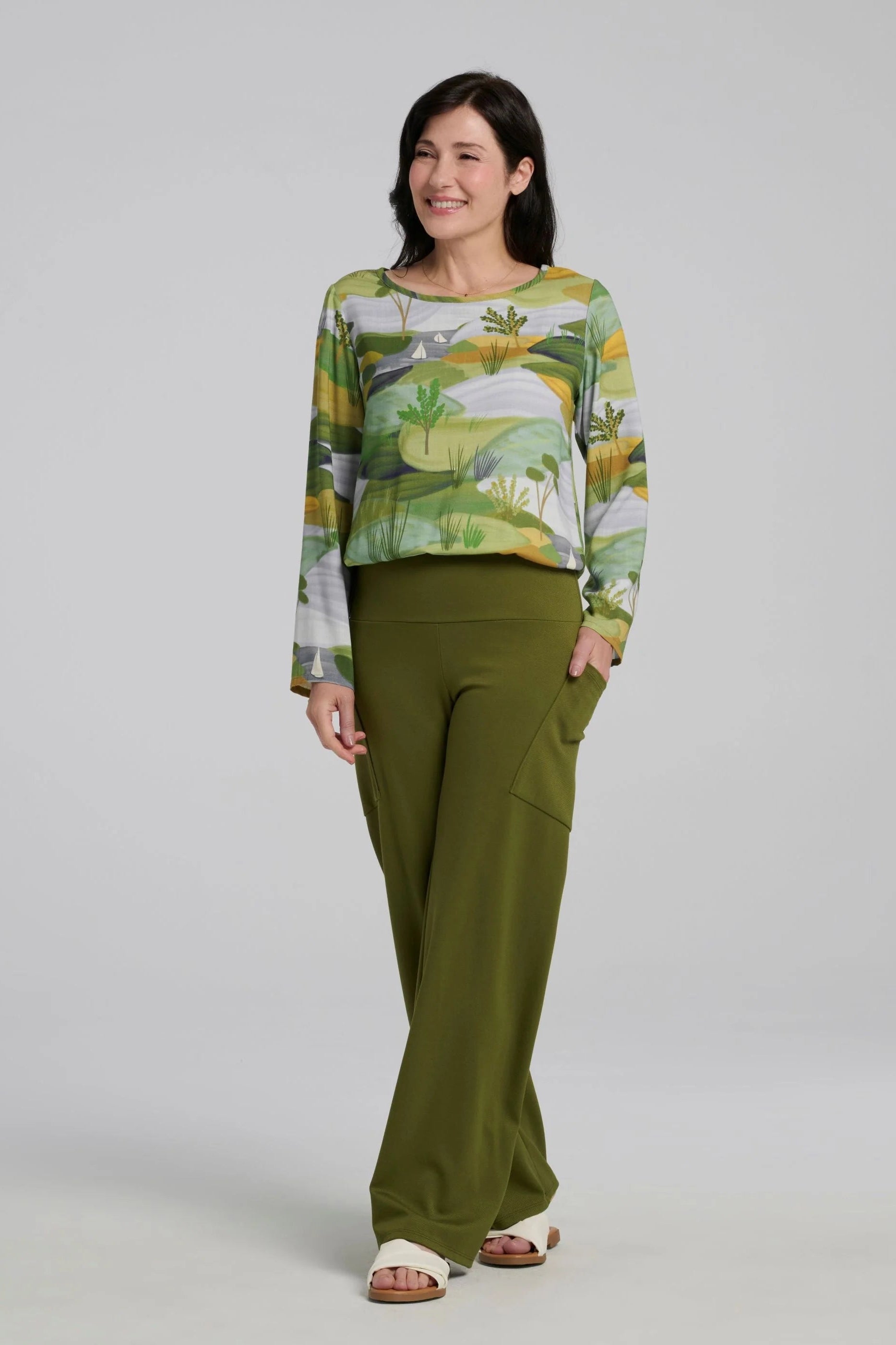 A woman wearing the spring Susan Pants by Kollontai in Granny Smith, wide leg pants with a pull-on power mesh lined waistband and angled patch pockets. She is wearing them with a patterned top and standing in front of a grey background. 