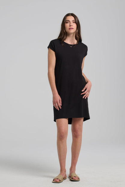 A woman wearing the Arwen Dress by Kollontai, a Black t-shirt dress with a round neck, short extended sleeves, a loose ft, and an above the knee hem. She is standing in front of a grey background. 