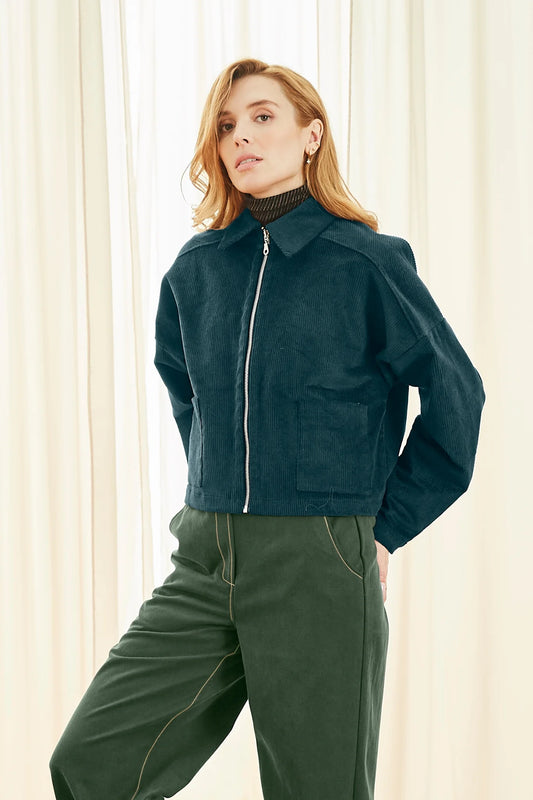 A woman wearing the Lennon Jacket by Melow in Petrole (Teal/Green), a short corduroy jacket with a blouse collar, zip front and patch pockets. She is wearing it with green pants and standing in front of a white curtain. 