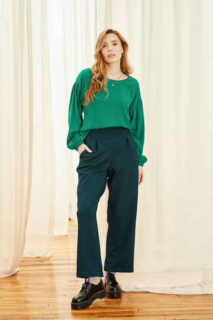 A woman wearing the Leo Pants by Melow in Petrole (Teal/Green), high-waisted full length pants with a wide pull-on waistband, straight legs, front pleats, and a diagonal stripe texture. She is wearing them with a green and  standing in front of white curtains.