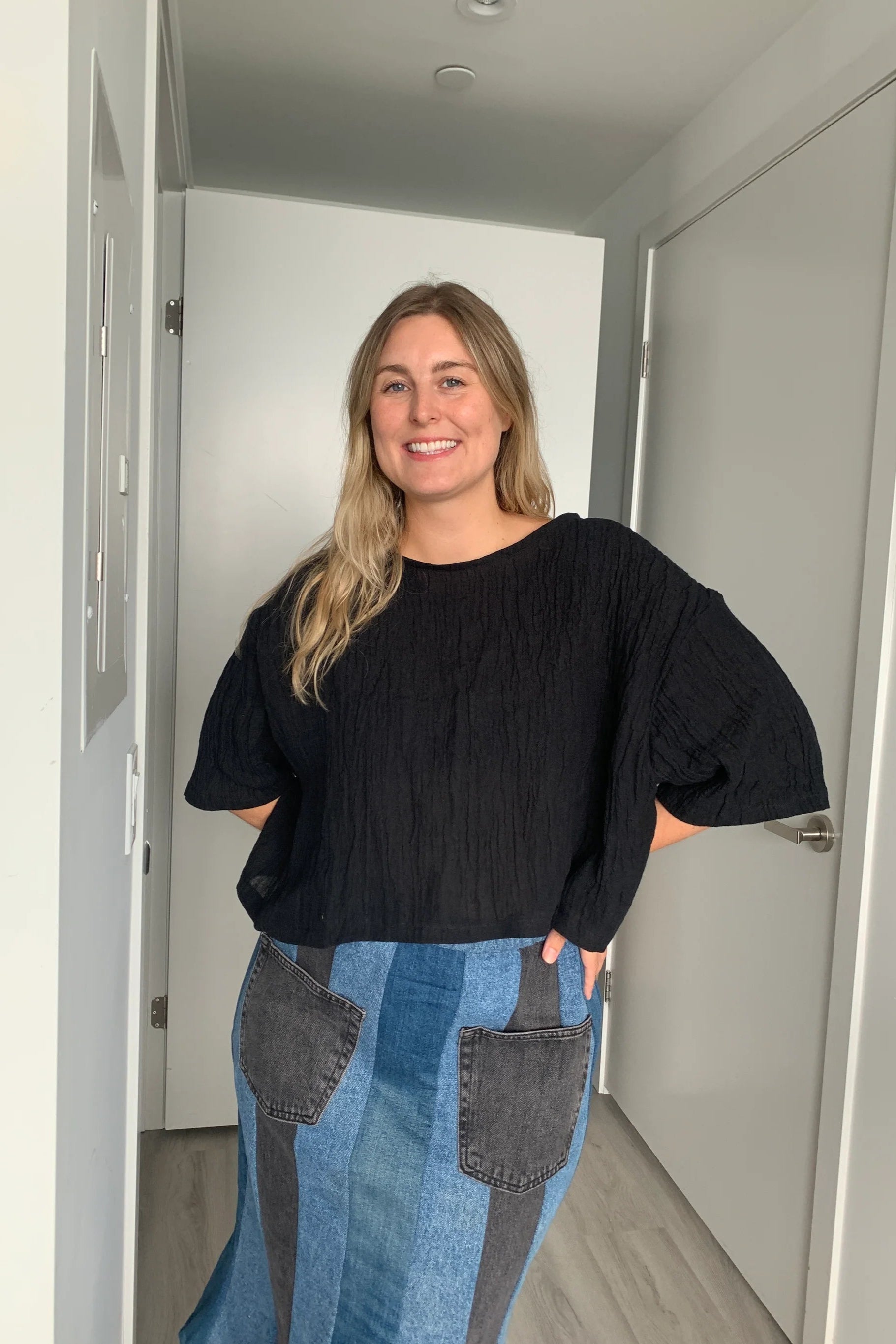  A woman wearing the Cropped Beach Tee by Lights of all in Black, a cropped linen t-shirt with 3/4 length drop sleeves and a boxy fit, and a slightly longer hem at the sides. She is wearing it with a patchwork denim skirt and standing in a white hallway. 