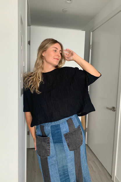  A woman wearing the Cropped Beach Tee by Lights of all in Black, a cropped linen t-shirt with 3/4 length drop sleeves and a boxy fit, and a slightly longer hem at the sides. She is wearing it with a patchwork denim skirt and standing in a white hallway. 