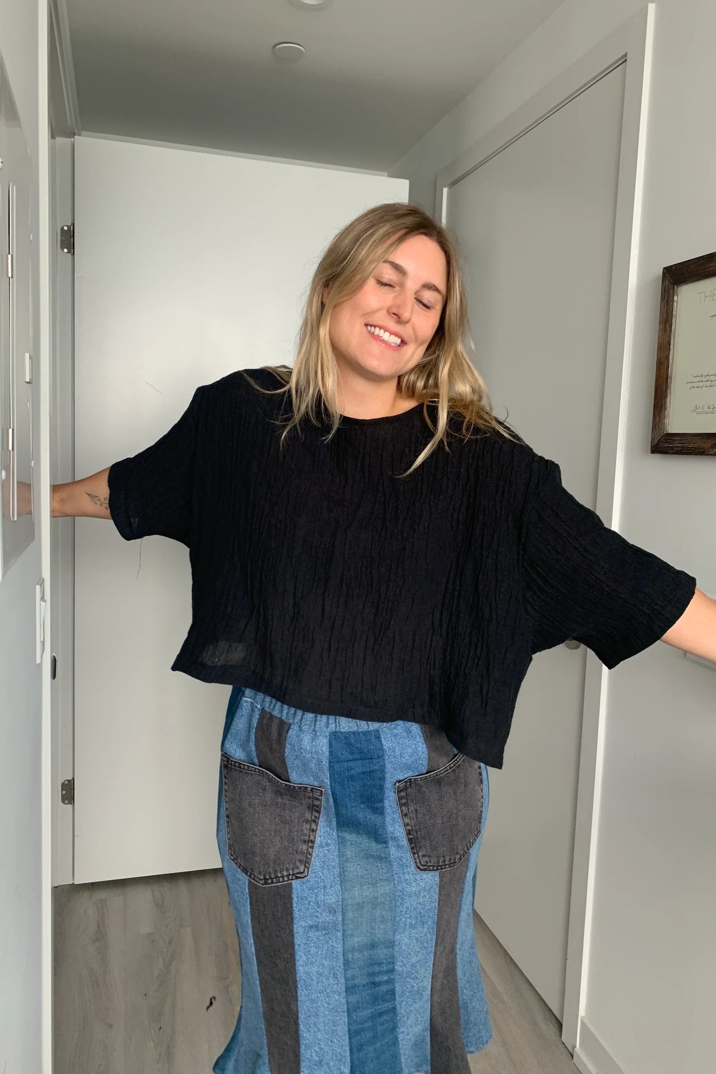  A woman wearing the Cropped Beach Tee by Lights of all in Black, a cropped linen t-shirt with 3/4 length drop sleeves and a boxy fit, and a slightly longer hem at the sides. She is wearing it with a patchwork denim skirt and standing in a white hallway. 
