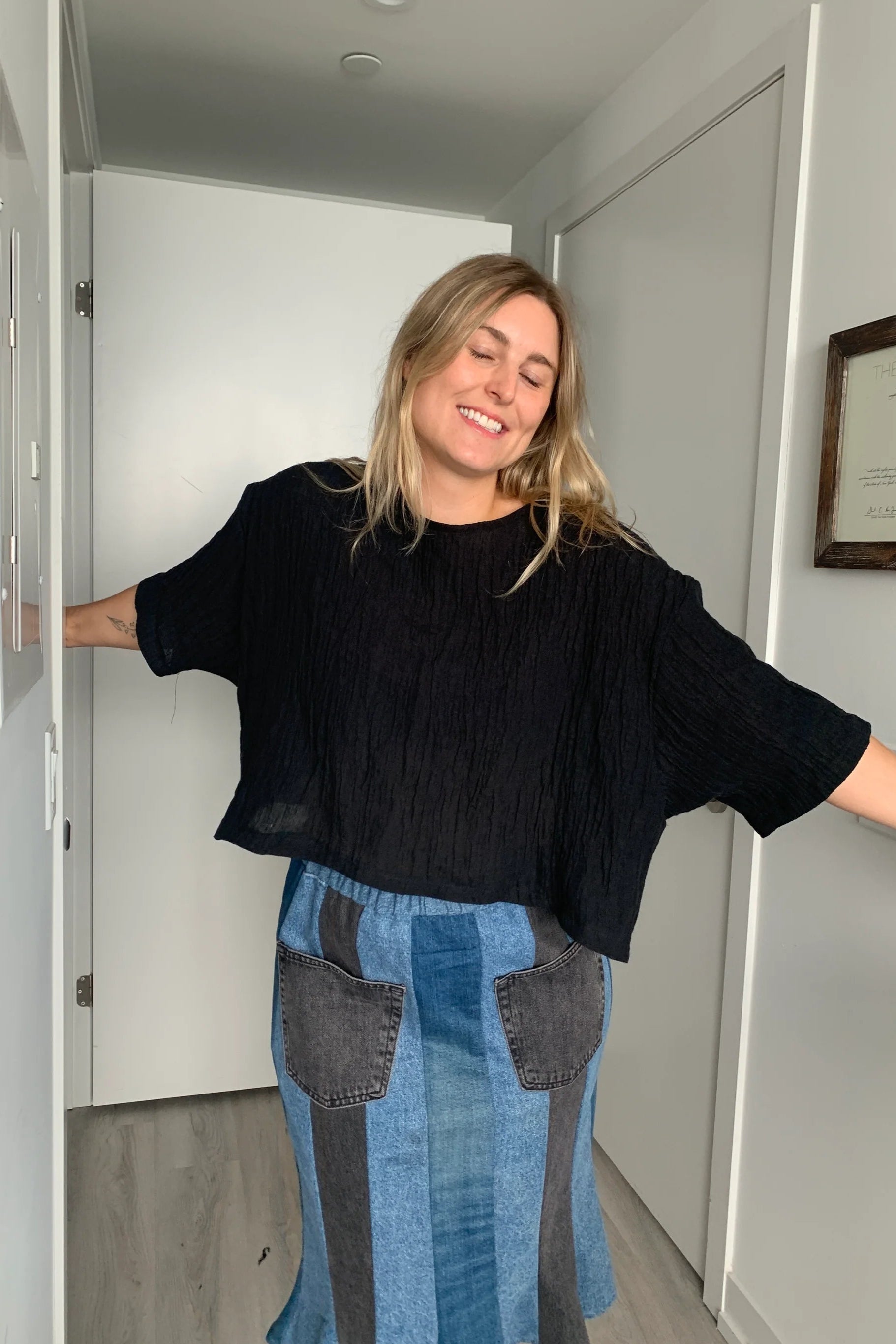  A woman wearing the Cropped Beach Tee by Lights of all in Black, a cropped linen t-shirt with 3/4 length drop sleeves and a boxy fit, and a slightly longer hem at the sides. She is wearing it with a patchwork denim skirt and standing in a white hallway. 