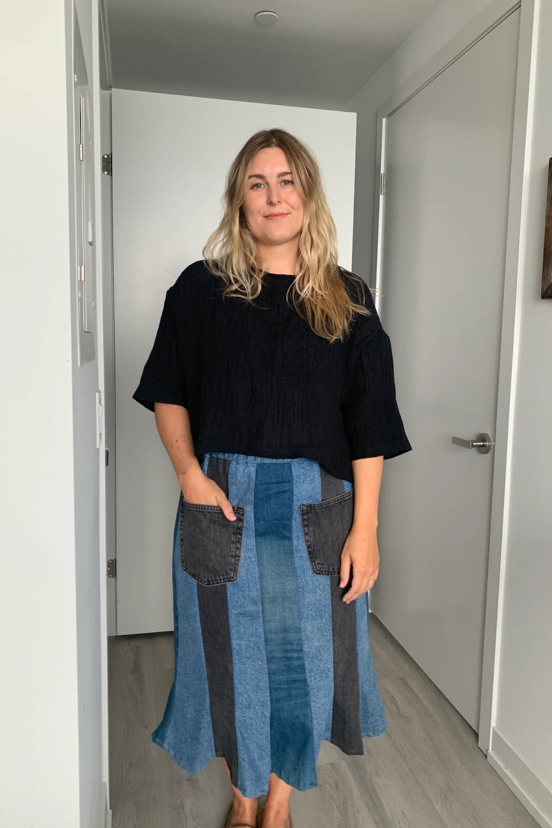  A woman wearing the Cropped Beach Tee by Lights of all in Black, a cropped linen t-shirt with 3/4 length drop sleeves and a boxy fit, and a slightly longer hem at the sides. She is wearing it with a patchwork denim skirt and standing in a white hallway. 