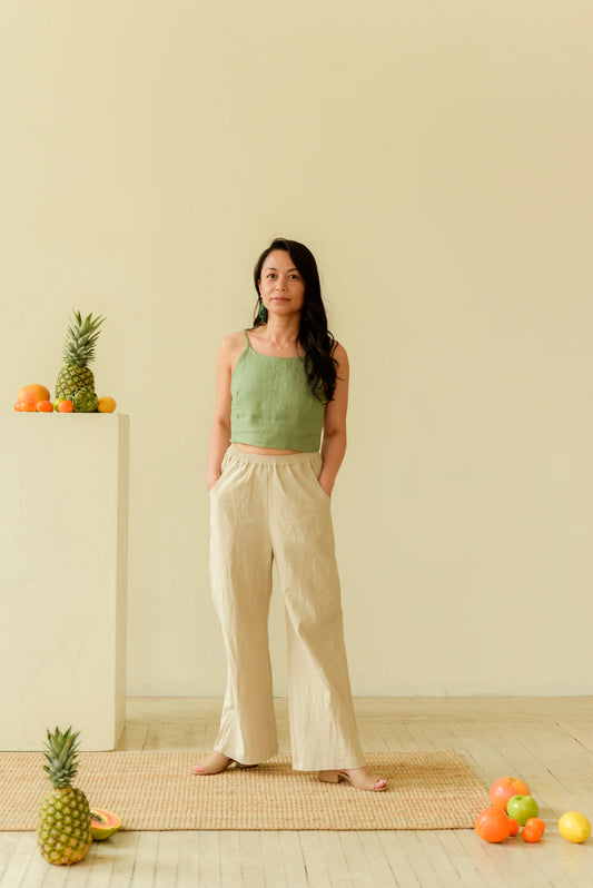 A woman wearing the Freedom Pants by Lights of All in Sand, featuring an elastic waist, wide legs, and pockets. She is wearing them with a green tank and standing in a beige room with fruit scattered around her. 