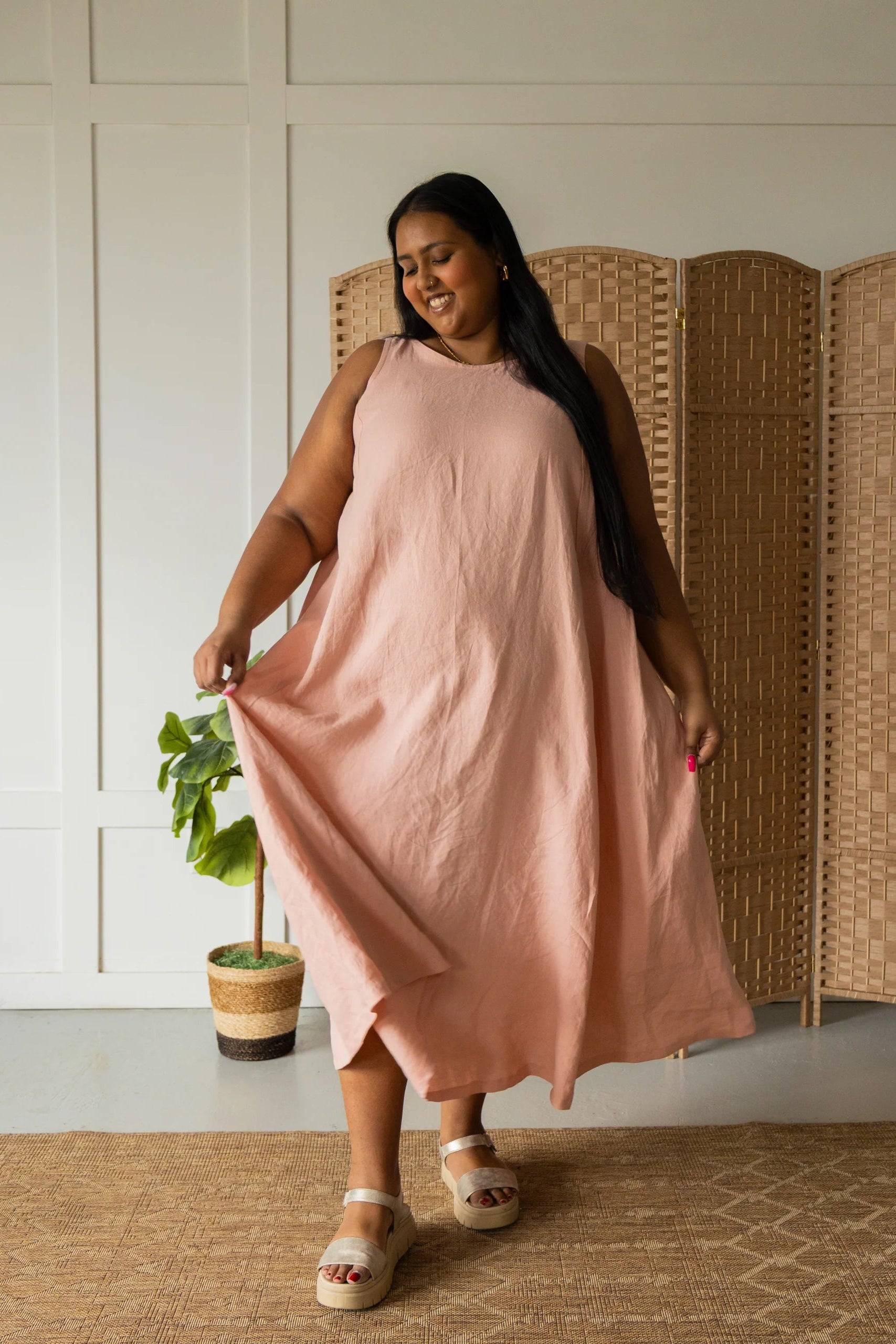 A woman wearing the Caroline Dress by Lights of All in Ballet Pink, a sleeveless dress linen maxi dress with a V-neck at the back. She is standing against a white wall with a woven screen behind her. 