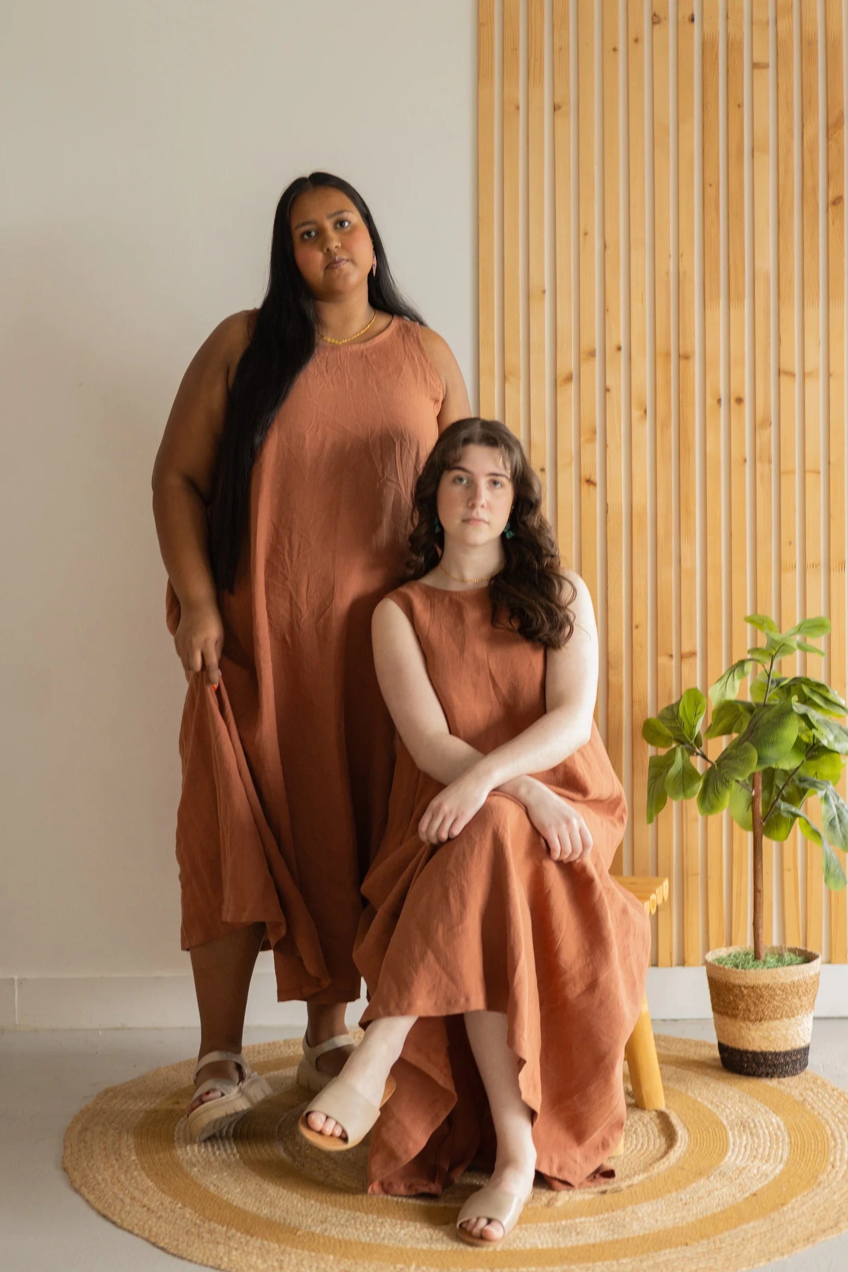 Two women wearing the Caroline Dress by Lights of All in Sedona, a sleeveless dress linen maxi dress with a V-neck at the back. One is seated on a stool and the other is standing, they are in front of a white and wood panelled wall. 