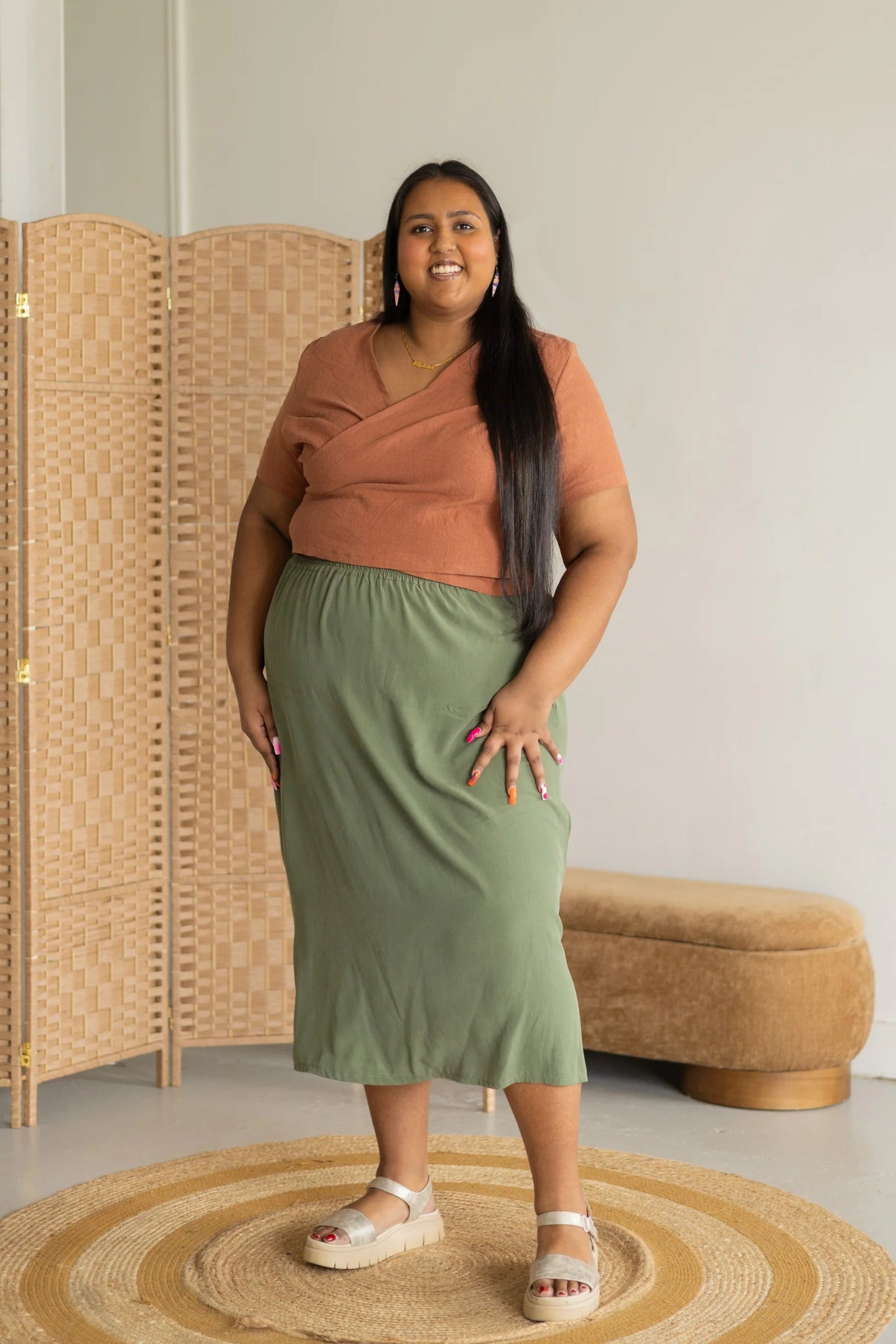 A woman wearing the Simone Skirt by Lights of All in Cactus, a sleek calf-length skirt with a flared hem and an elastic waist. She is wearing it with the Stella Top in Sedona, and standing by a woven screen.