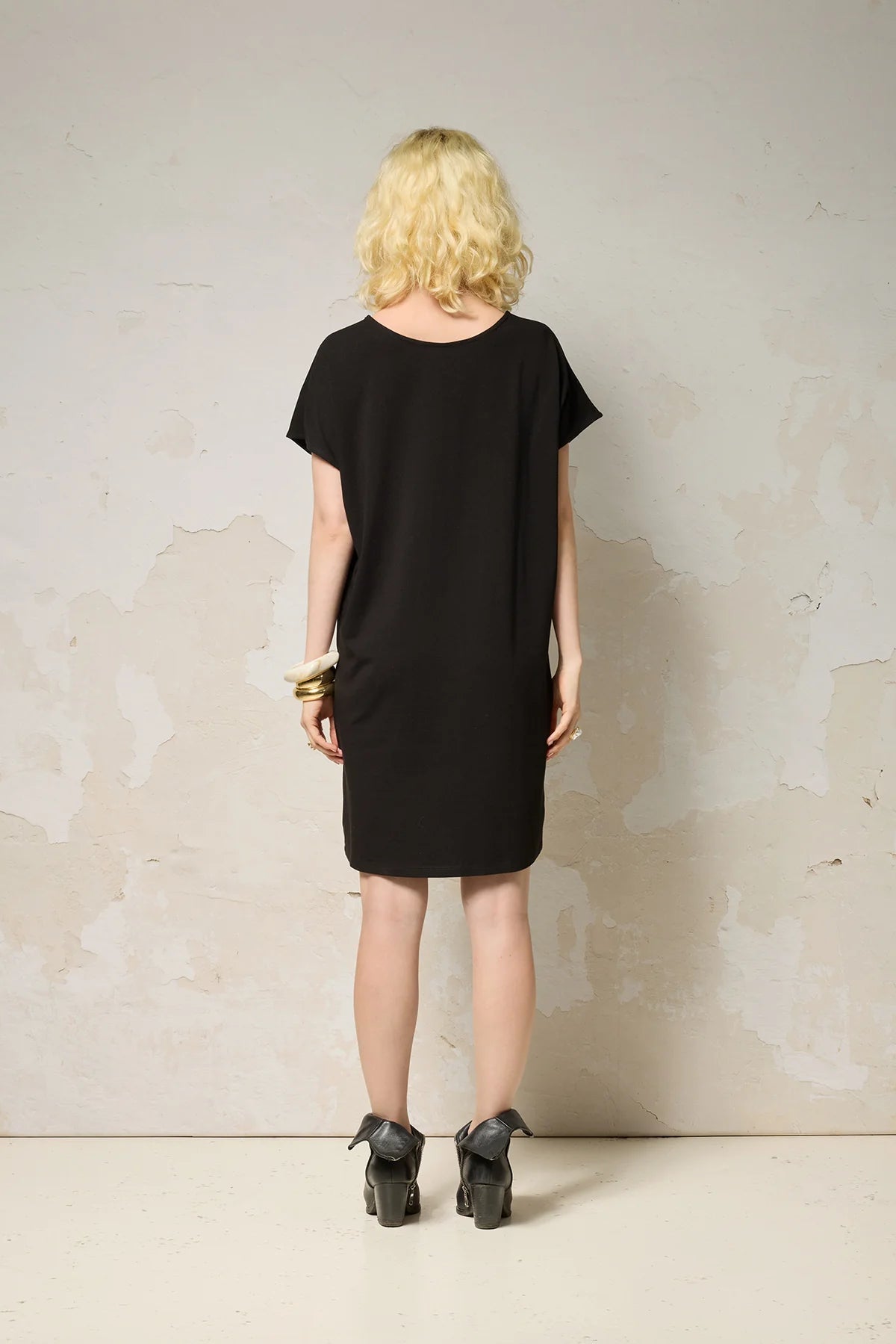Back view of a woman wearing the Lyssa Dress by Cokluch in Black, a short dress with a fringed v-panel at the cheset, short sleeves, and a relaxed fit. She is standing against a textured wall.