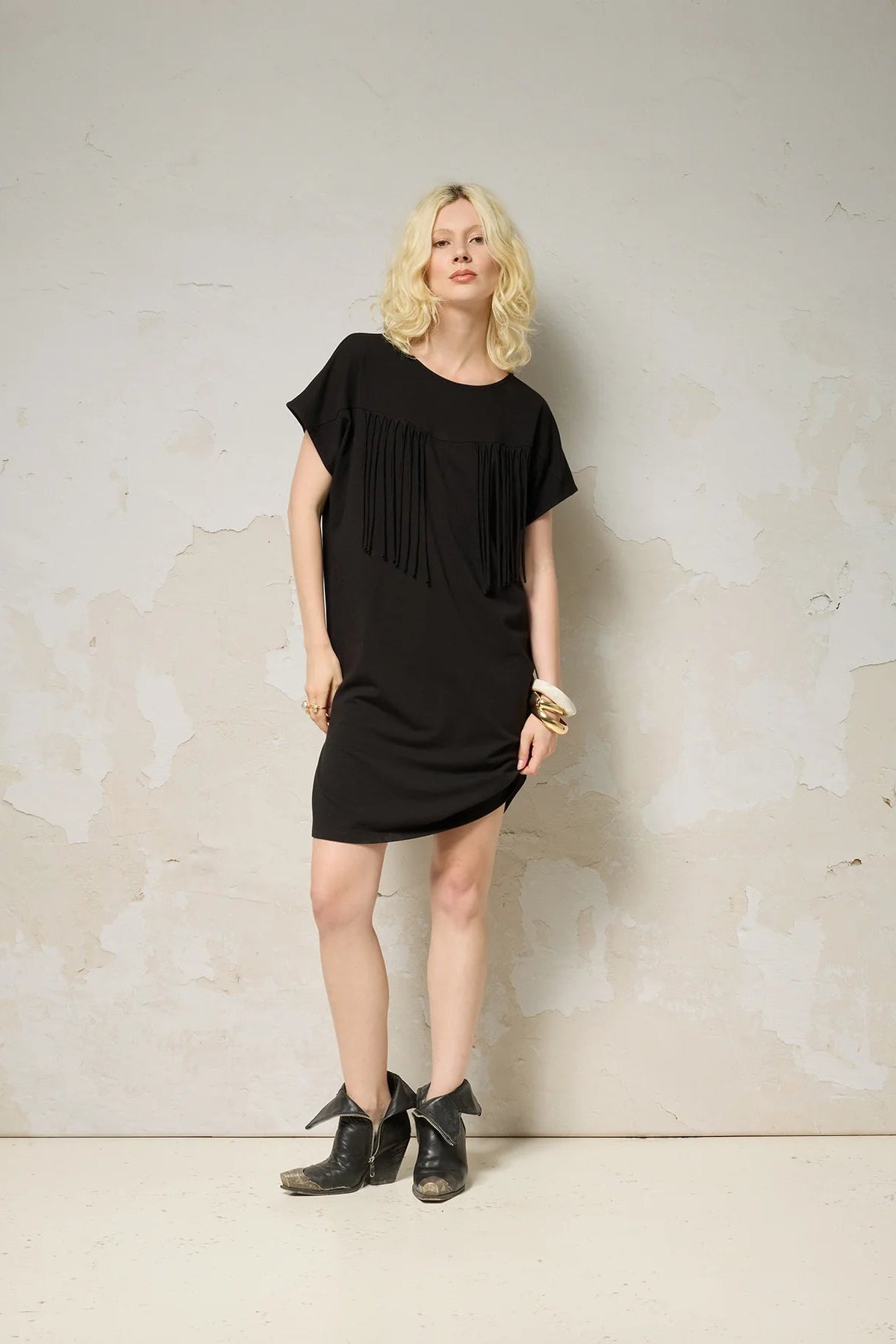 A woman wearing the Lyssa Dress by Cokluch in Black, a short dress with a fringed v-panel at the cheset, short sleeves, and a relaxed fit. She is standing against a textured wall.