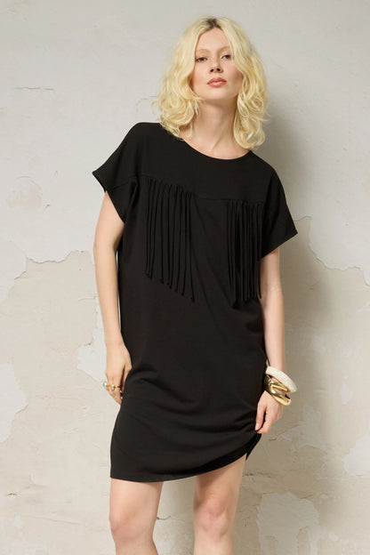 3/4 view of a woman wearing the Lyssa Dress by Cokluch in Black, a short dress with a fringed v-panel at the cheset, short sleeves, and a relaxed fit. She is standing against a textured wall.