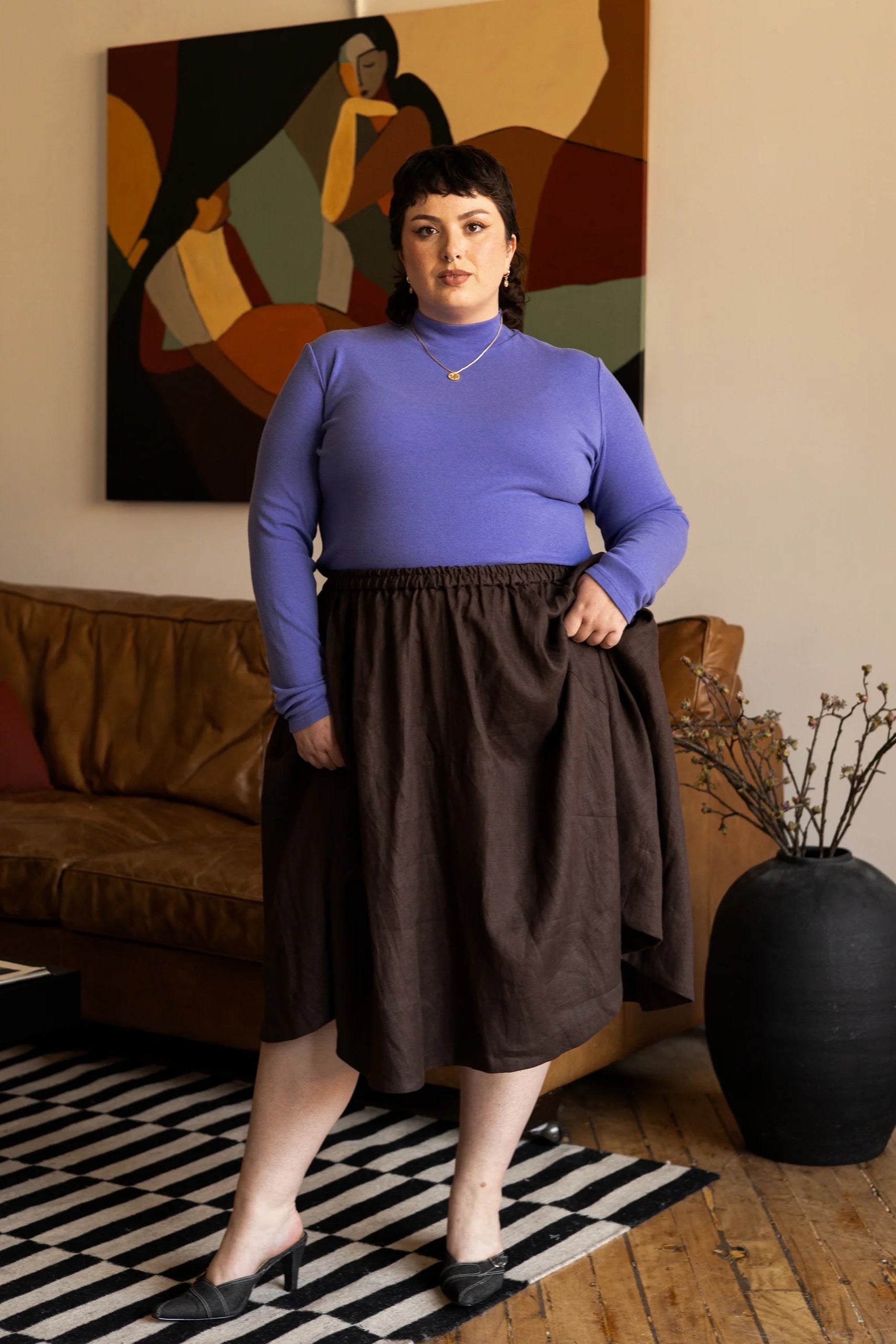 Woman wearing the Tuesday Skirt by Lights of All in Cocoa with the Essential turtleneck in Violet standing in a living room with abstract art on the wall.