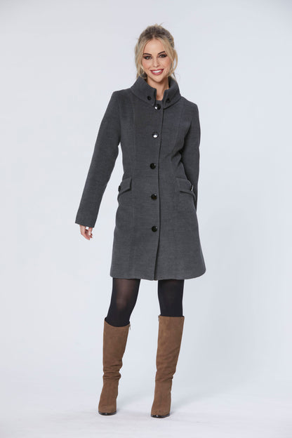 A woman wearing the Josephine Jacket by Luc Fontaine in Grey, a mid-thigh length coat featuring a high collar with button details, a button front, and flap pockets with buttons. She is standing in front of a white background 