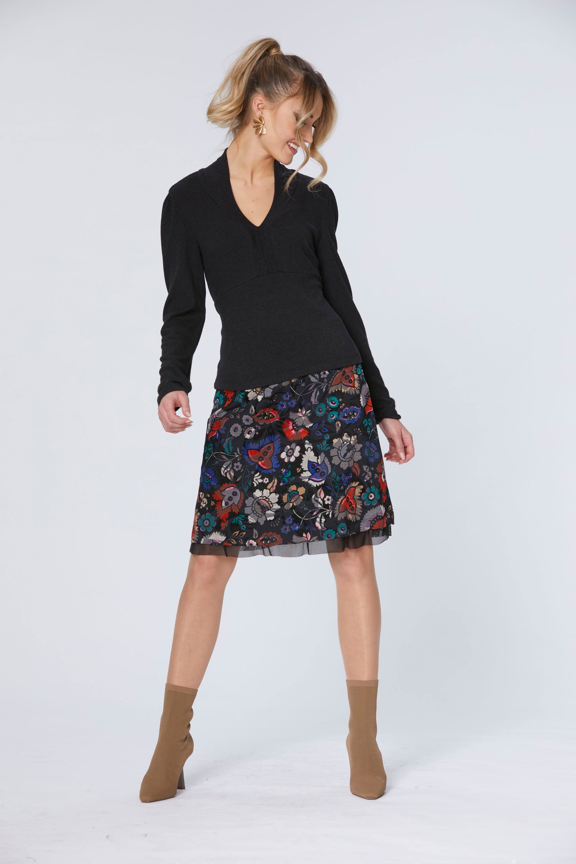 A woman wearing the Slight Top by Luc Fontaine in Black, with a v-neck, panels around the neckline, a under-bust seam, and a long sleeves, with the Mistery Skirt by Luc Fontaine in Multicolour Print. She is standing in front of a white background. 