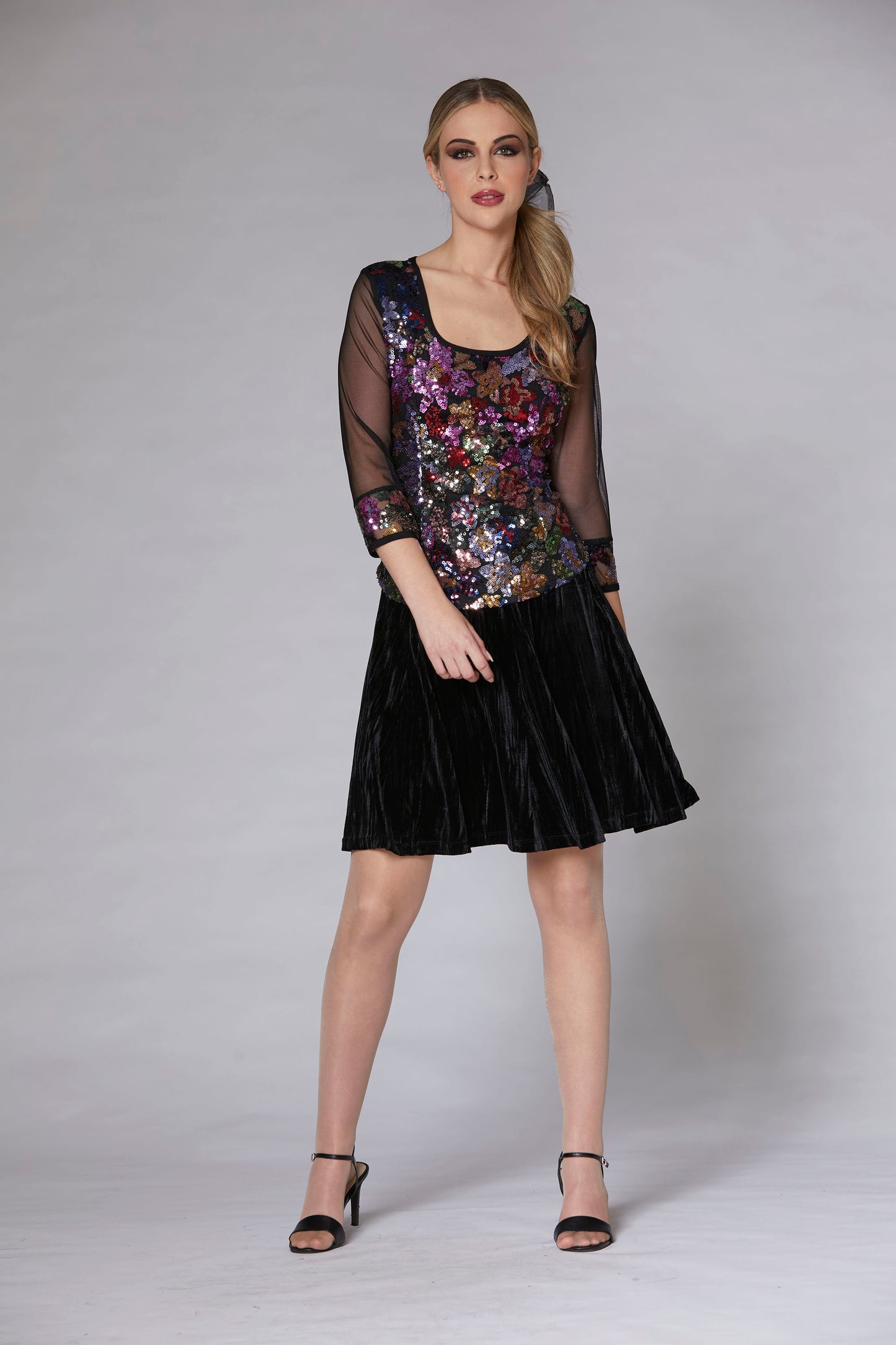 A woman wearing the Glam sequin top by Luc Fontaine, a floral sequin top with sheer black sleeves with sequins at the cuffs. She is wearing it with the Lustra Skirt in black velvet  and is standing in front of a grey background. 