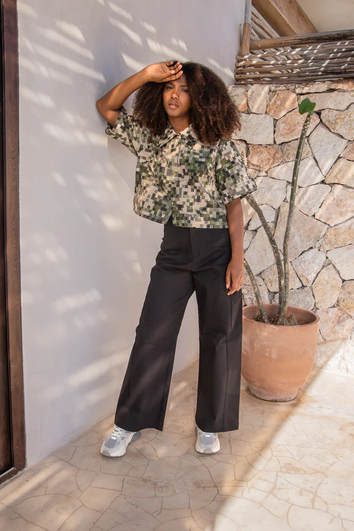 A woman wearing the Almeria Top by MAS in Camo, a slightly cropped button up short sleeved shirt with a large collar and a pixellated print in green and earth tones. She's wearing it with the Vienna Pants in Black, and standing outside by a stone wall. 