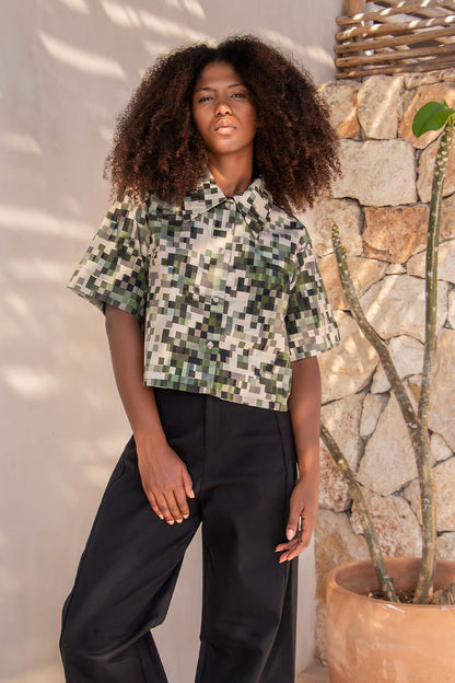 A woman wearing the Almeria Top by MAS in Camo, a slightly cropped button up short sleeved shirt with a large collar and a pixellated print in green and earth tones. She's wearing it with the Vienna Pants in Black, and standing outside by a stone wall. 
